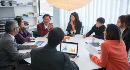 A group of people are sitting around a table having a meeting. There are five men and two women in the group. They are all wearing business suits. The table is covered in papers and there is a laptop on the table. The people are all looking at the man at the head of the table. He is speaking and gesturing with his hands. The people are all listening attentively. It appears that they are in the middle of a serious discussion.