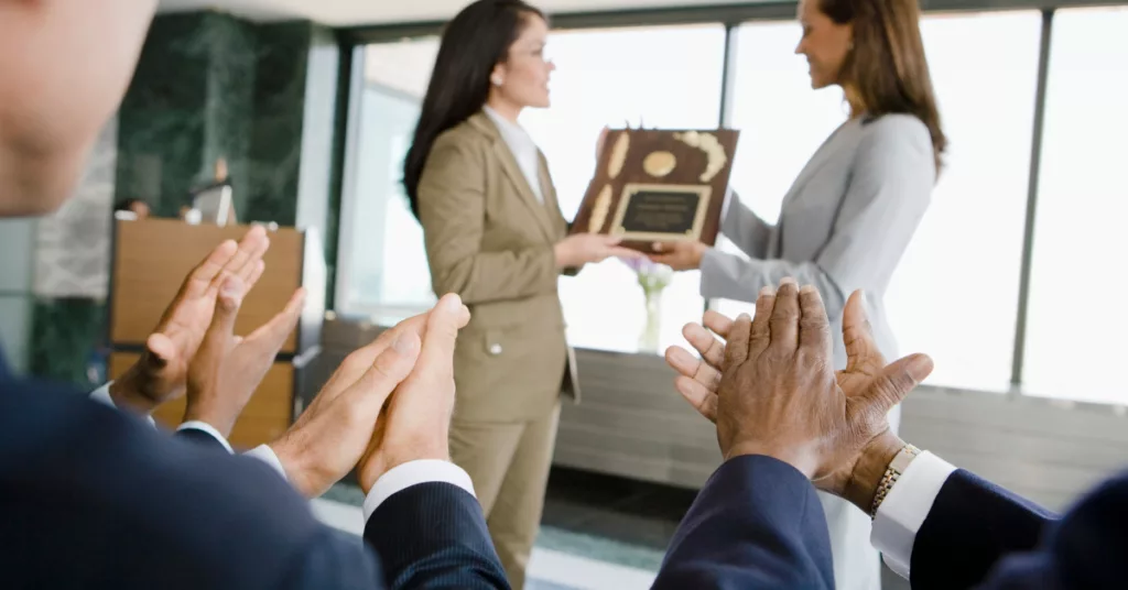 How Instant Gratification Affect the Way We Work. Photo of a woman receiving an award from her boss with her coworkers clapping. 