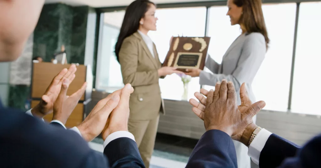 How Instant Gratification Affect the Way We Work. Photo of a woman receiving an award from her boss with her coworkers clapping.