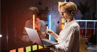 A woman in a headset is standing in front of a glass board and holding a laptop and marker. She is looking at the laptop and writing on the glass board. She is wearing a white blouse and has short blonde hair.